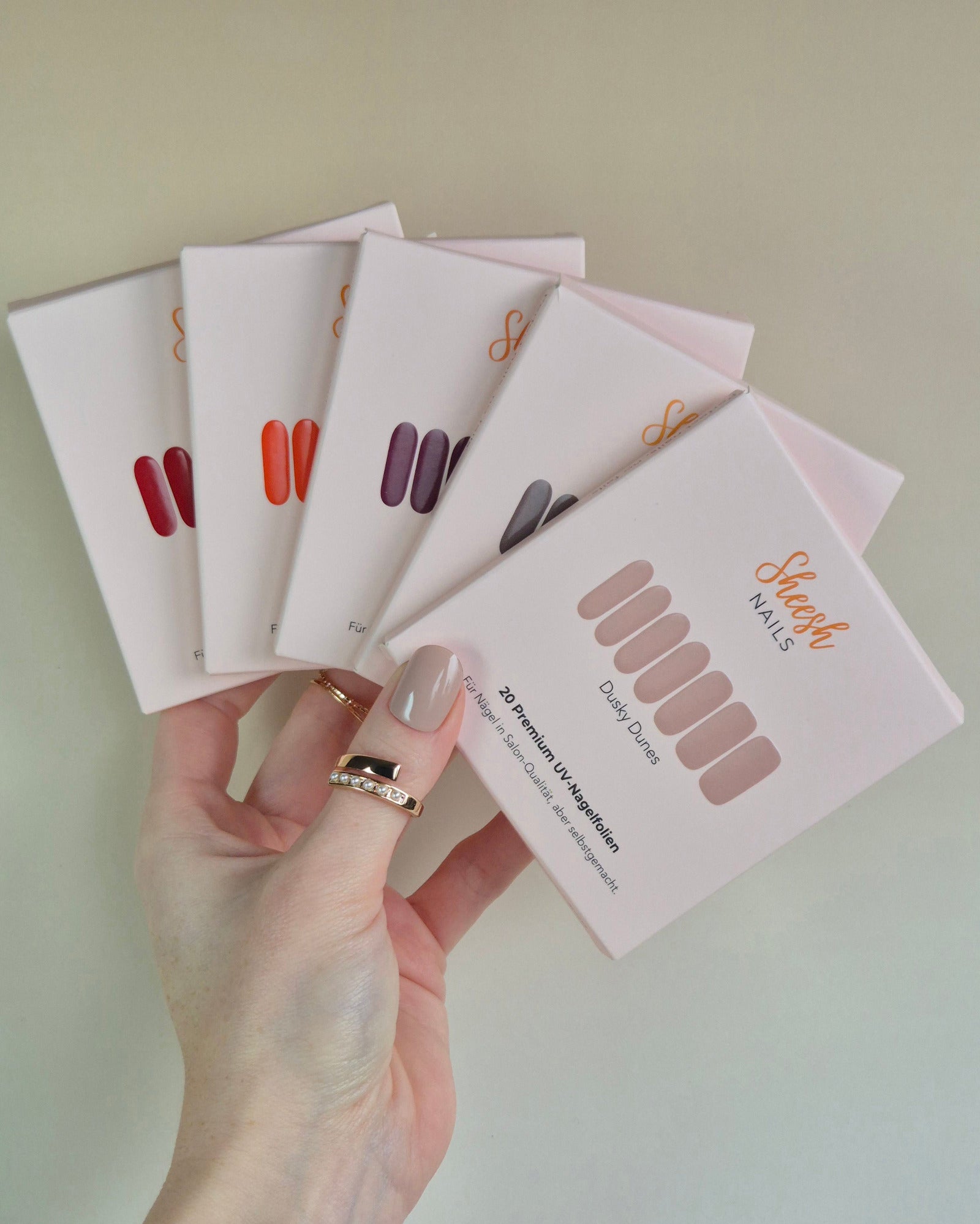 Sheesh UV-Nagelfolien 5er Set – Hand hält fünf verschiedene Sheesh Nagelfolien-Verpackungen in Salonqualität für individuelle Maniküre.