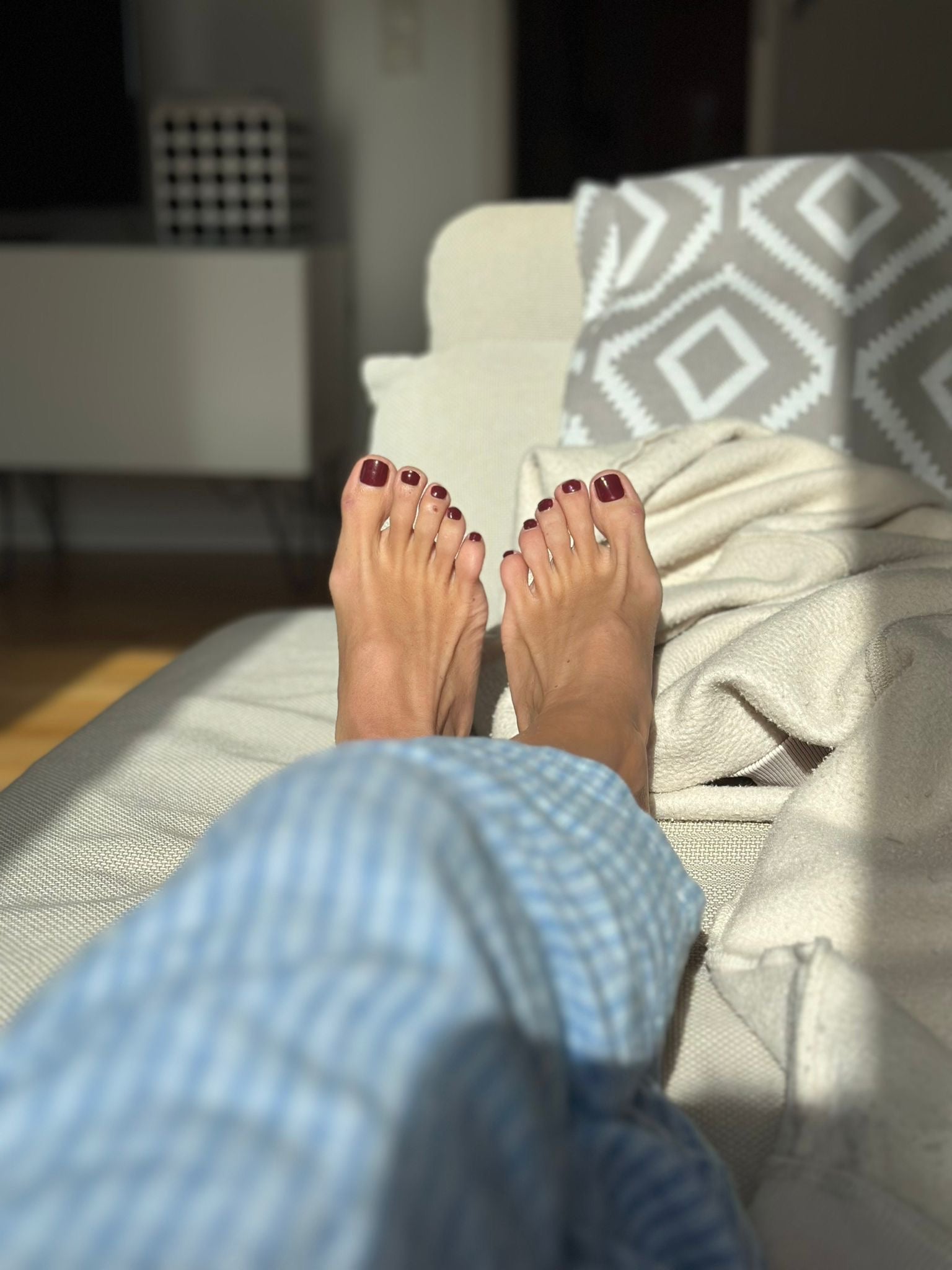 Füße mit dunkelvioletten Sheesh Nails UV-Nagelfolien in der Farbe „LA by Night“, im Sonnenlicht auf dem Sofa. Pediküre mit UV-Folien für langanhaltenden Glanz und eleganten Look, perfekt für entspannte Selfcare-Momente zuhause.