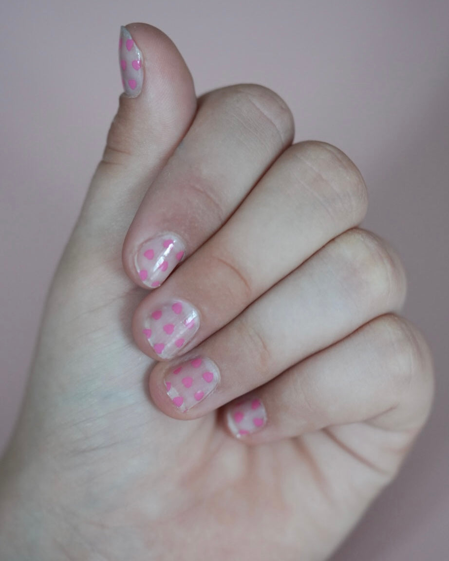 Kinderhand mit rosa Sheesh Nails Kinderfolie „Love“. Die Nagelfolien zeigen kleine, pinke Herzmotive auf transparentem Untergrund und verleihen einen verspielten, liebevollen Look – perfekt für Kinder, die zarte Farben und kreative, herzliche Designs lieben.