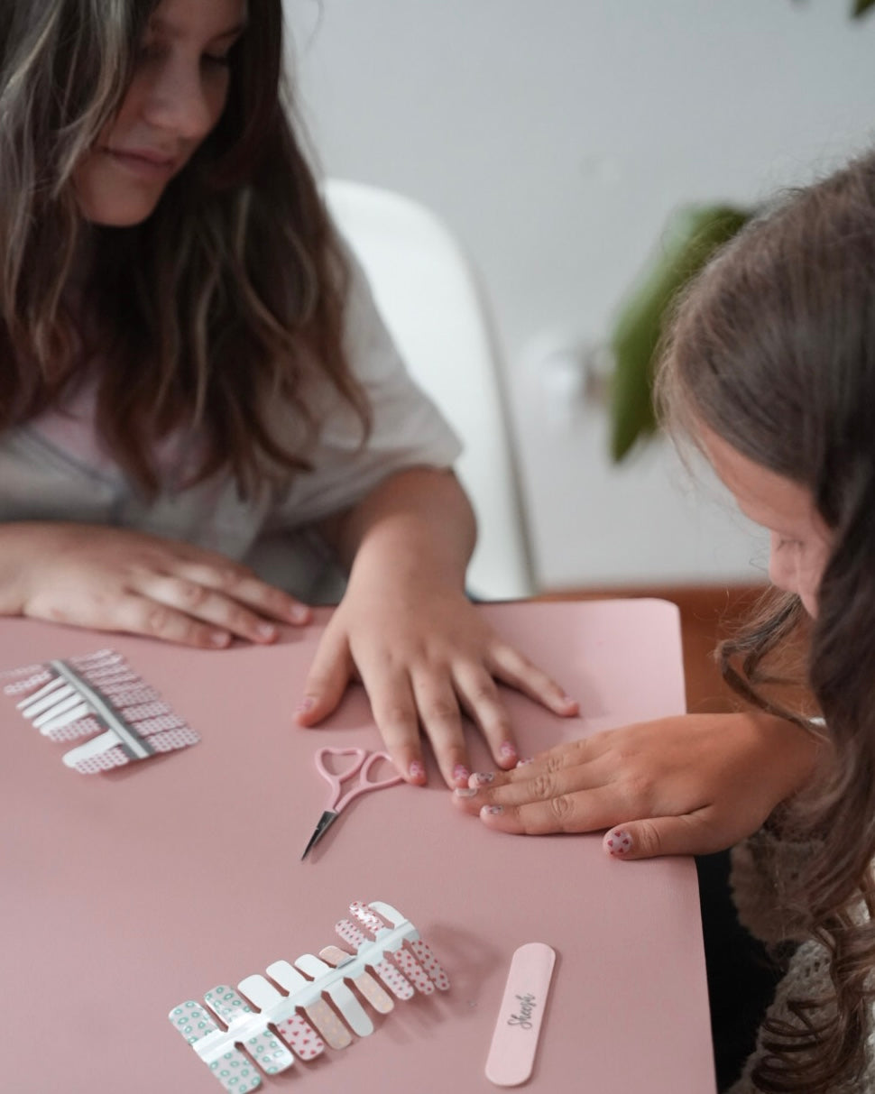 Zwei Kinder sitzen gemeinsam am Tisch und bereiten Sheesh Nails Kinderfolien „Süße Früchtchen“ vor. Auf dem rosa Untergrund liegen bunte Nagelfolien, Schere und Feile. Die Szene vermittelt spielerische Freude am Basteln und kreativen Gestalten – ideal für fröhliche Kinder, die Spaß an farbenfrohen Nageldesigns haben.
