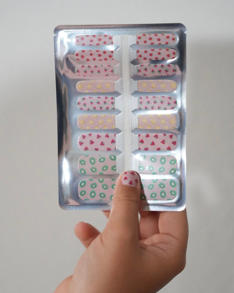 Kinderhand hält ein Set der Sheesh Nails Kinderfolie „Süße Früchtchen“. Die bunten Nagelfolien zeigen kleine Früchtemotive wie Erdbeeren, Zitronen und Kiwis auf transparentem Untergrund. Das verspielte Design steht für Sommer, Spaß und kreative Maniküren – ideal für Kinder, die fröhliche, farbenfrohe Styles lieben.