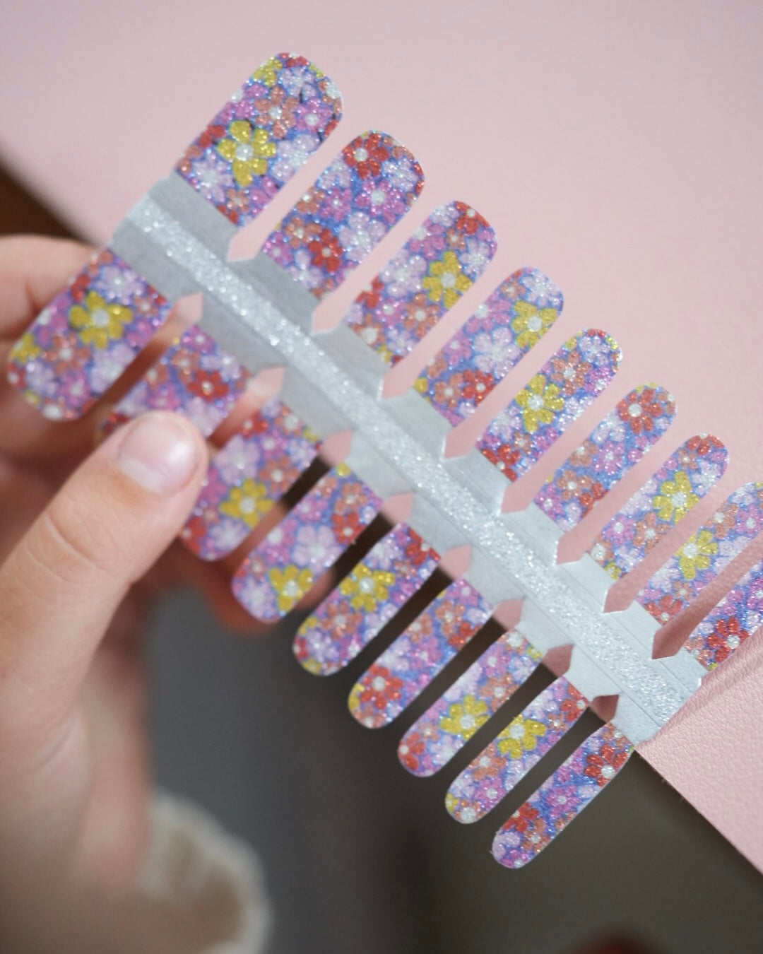 Hand hält ein Set der Sheesh Nails Kinderfolie „Flower Power“. Die bunten Nagelfolien zeigen ein fröhliches Blumenmuster in Lila-, Rosa- und Gelbtönen mit glitzerndem Effekt – kindgerechtes, verspieltes Design für kreative kleine Nagelfans und farbenfrohe Maniküre.