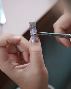 Kinderhand schneidet mit einer kleinen Schere ein Stück der Sheesh Nails Kinderfolie „Flower Power“ zurecht. Die Nagelfolie zeigt bunte Blumenmotive in Lila-, Rosa- und Gelbtönen mit Glitzereffekt. Das Bild vermittelt kreatives Basteln und Spaß an kindgerechter Nailart – ideal für kleine Nagelfans, die farbenfrohe, verspielte Designs lieben.