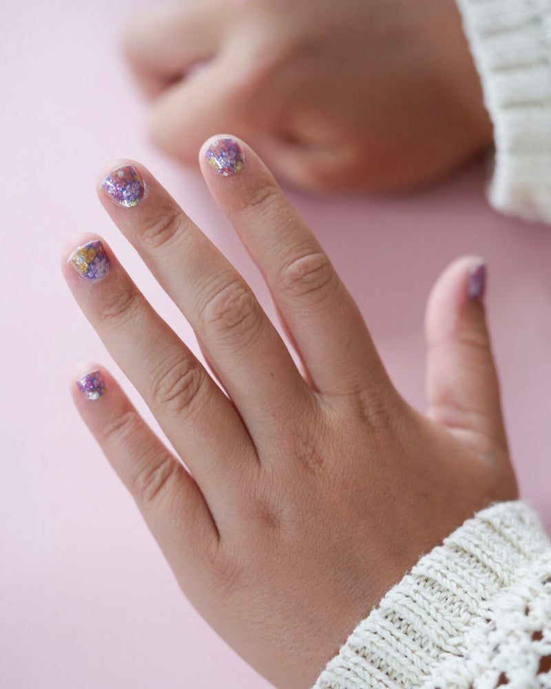 Kinderhand mit bunten, glitzernden Nagelfolien zeigt die Sheesh Nails Kinderfolie „Flower Power“. Das fröhliche Design mit kleinen Blumenmotiven in Lila-, Rosa- und Gelbtönen sorgt für einen verspielten, kreativen Look – perfekt für Kinder, die Spaß an farbenfroher, glänzender Maniküre haben.