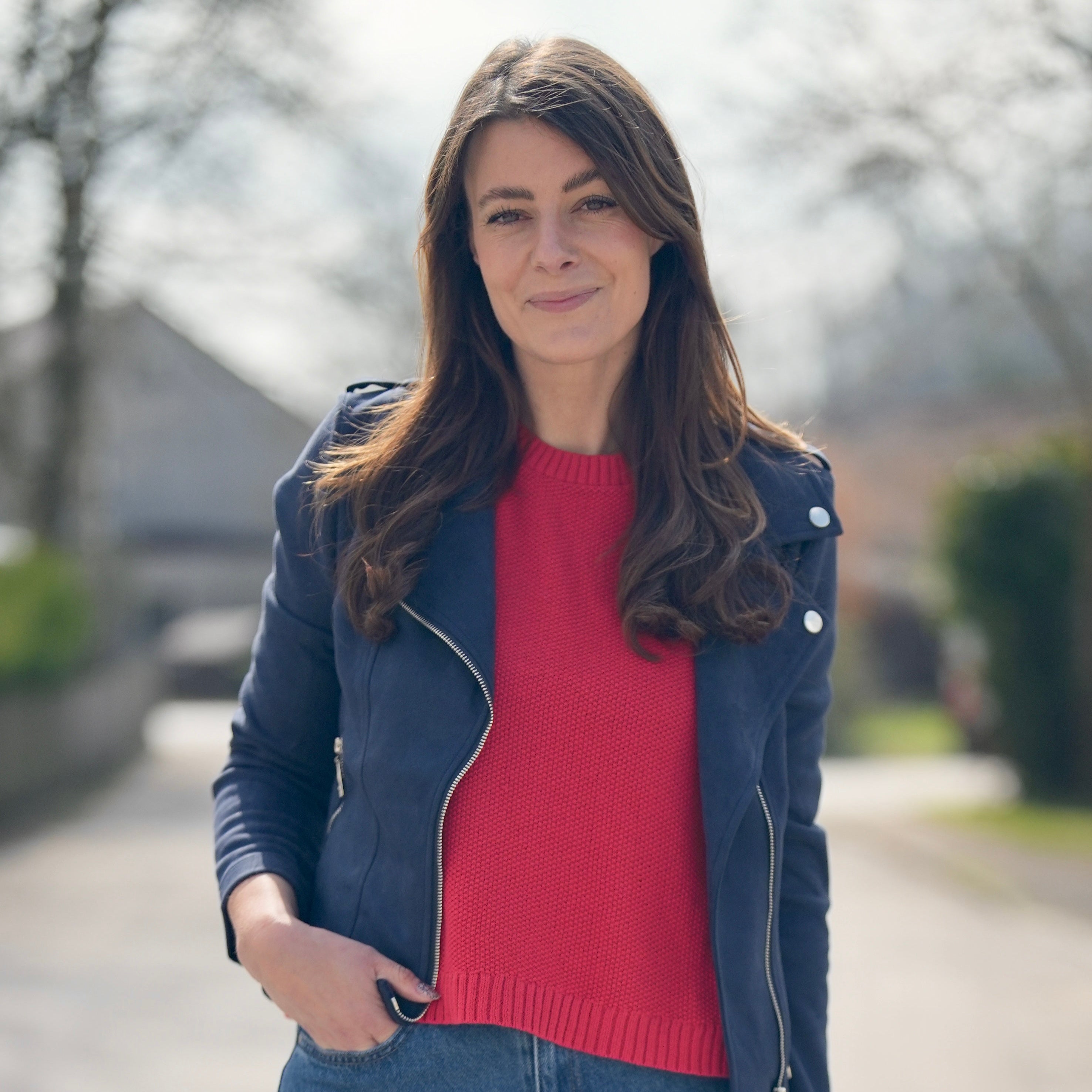 Porträtaufnahme einer Frau im Freien. Sie trägt eine blaue Jacke, ein rotes Oberteil und Jeans. Der Hintergrund ist leicht unscharf und zeigt eine sonnige Straße mit Bäumen und Gebäuden.