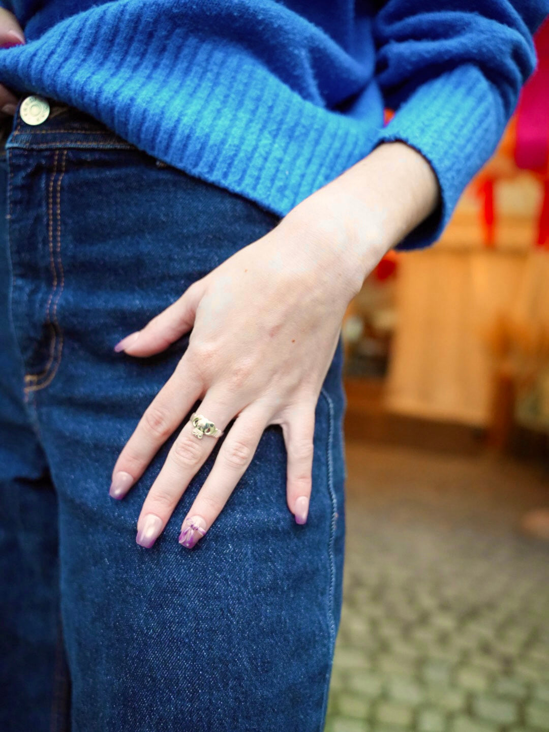 Sheesh Nails Press-on Nails in zartem Rosa-Lila-Ton mit dezentem Glanz und floralem Design. Stilvolle, wiederverwendbare künstliche Fingernägel für einen modernen Alltagslook. Hand mit femininer Maniküre, kombiniert mit Jeans und blauem Pullover – elegantes, trendbewusstes Styling für jeden Anlass.