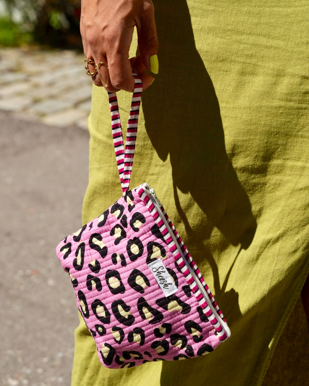Sheesh Nails Beauty-Bag – pinke Kosmetiktasche mit Leopardenmuster, gehalten von einer Frau in olivgrünem Sommeroutfit, sonniger Tag, moderner Lifestyle, Nagelfolien-Accessoire für unterwegs.