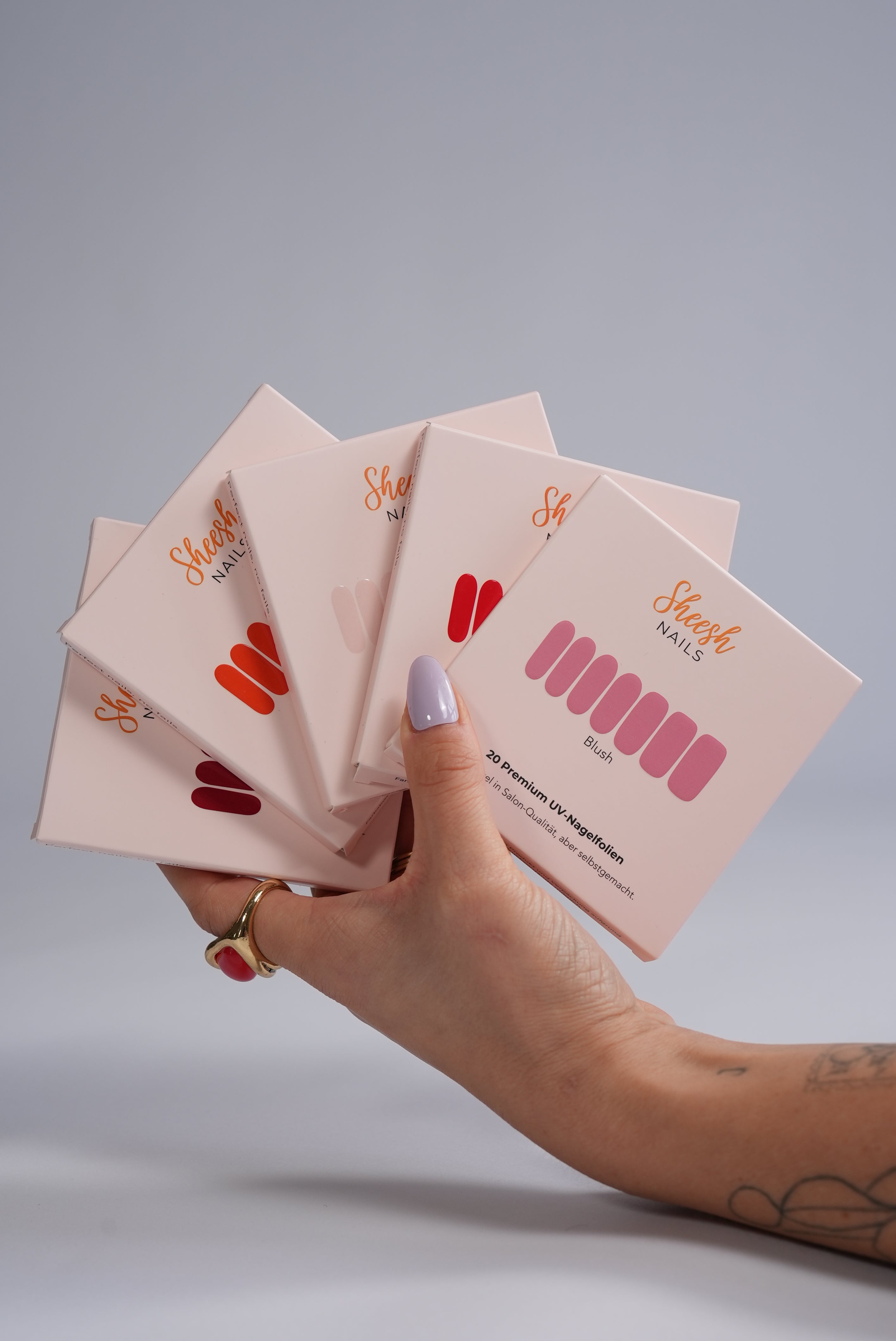 Sheesh UV-Nagelfolien 5er Set – Hand hält fünf verschiedene Sheesh Nagelfolien-Verpackungen in Salonqualität für individuelle Maniküre.