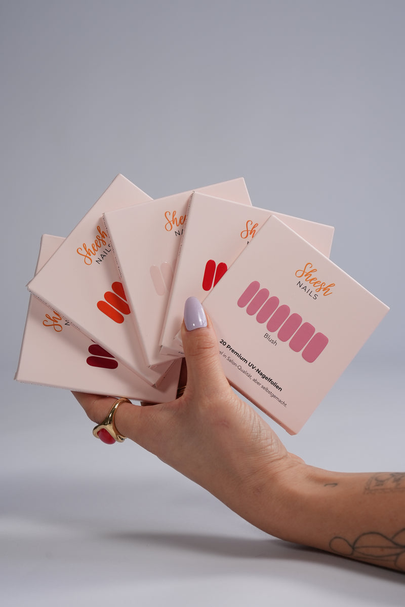 Sheesh UV-Nagelfolien 5er Set – Hand hält fünf verschiedene Sheesh Nagelfolien-Verpackungen in Salonqualität für individuelle Maniküre.