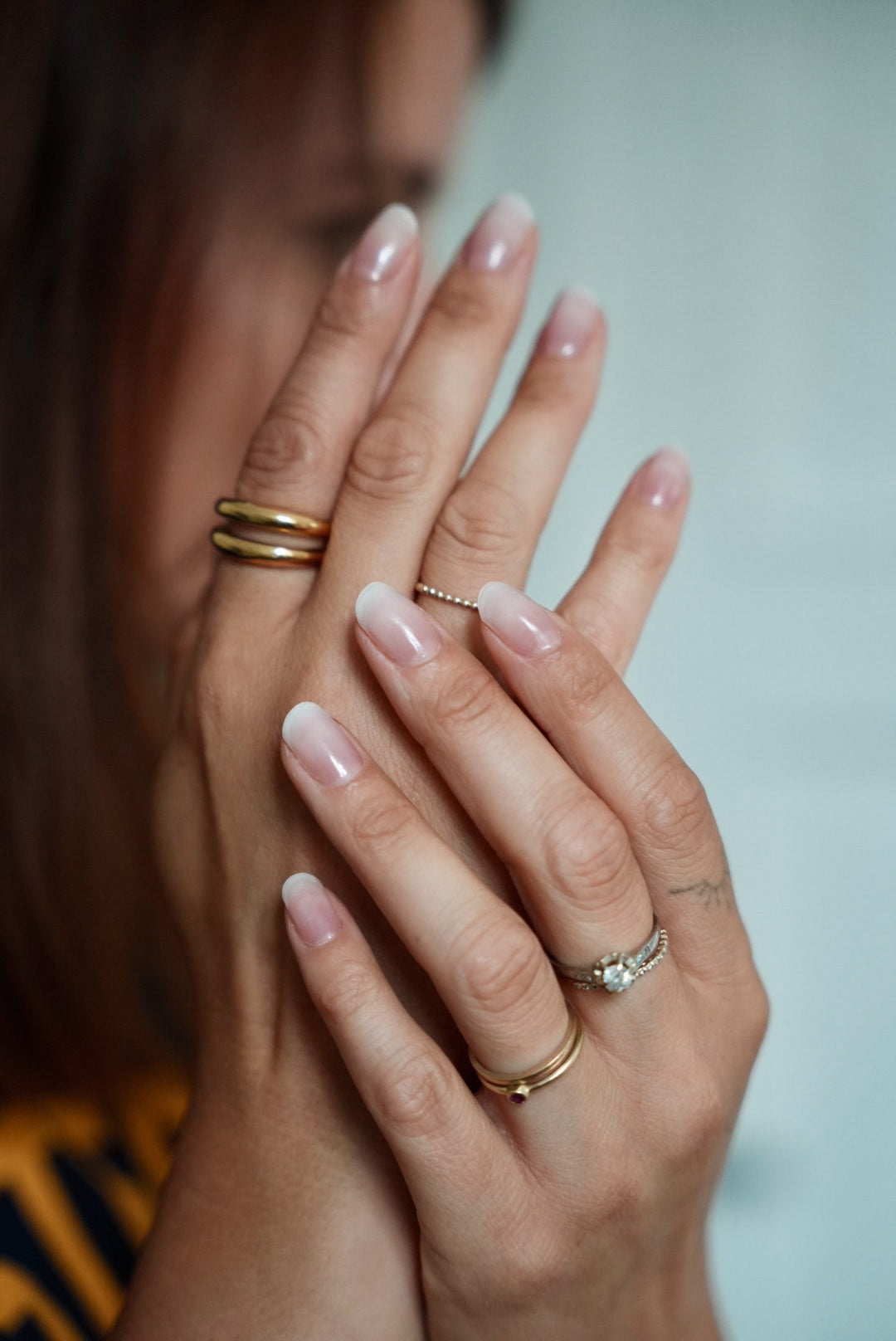 Nahaufnahme von Händen mit gepflegten Nägeln in natürlichem Glanz. Die Finger sind leicht ineinander verschränkt, mehrere goldene und silberne Ringe schmücken die Hände. Der Hintergrund ist weich unscharf, wodurch der Fokus auf den Nägeln und Schmuckdetails liegt.