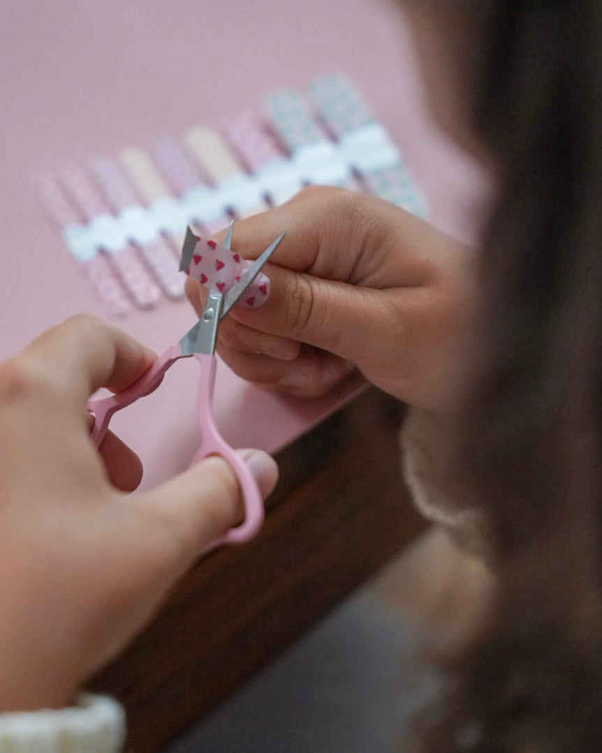 Kinderhand schneidet mit einer rosafarbenen Schere ein Stück der Sheesh Nails Kinderfolie „Love“ aus. Die Nagelfolien zeigen kleine rosa Herzen auf transparentem Untergrund. Die Szene vermittelt spielerische Kreativität und Freude – ideal für Kinder, die gerne basteln und liebevolle, farbenfrohe Designs mögen.