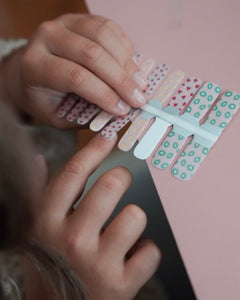 Kinderhand hält ein Set der Sheesh Nails Kinderfolie „Freche Früchtchen“. Die bunten Nagelfolien zeigen fröhliche Fruchtmotive wie Erdbeeren, Zitronen und Kiwis auf transparentem Untergrund. Das verspielte Design steht für Sommer, Spaß und kreative Kinder-Maniküren – ideal für kleine Nagelfans, die farbenfrohe Styles lieben.