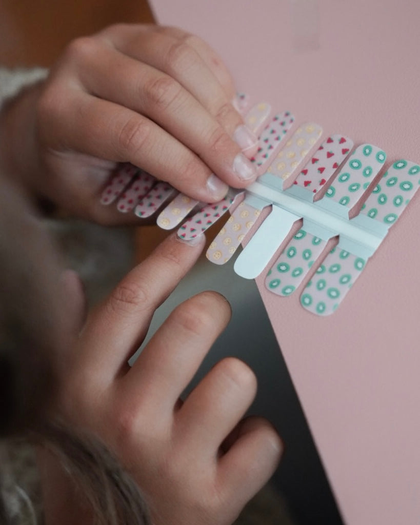 Kinderhand hält ein Set der Sheesh Nails Kinderfolie „Freche Früchtchen“. Die bunten Nagelfolien zeigen fröhliche Fruchtmotive wie Erdbeeren, Zitronen und Kiwis auf transparentem Untergrund. Das verspielte Design steht für Sommer, Spaß und kreative Kinder-Maniküren – ideal für kleine Nagelfans, die farbenfrohe Styles lieben.