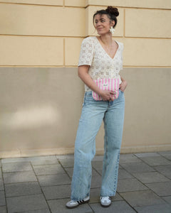 Frau mit lässigem Sommer-Outfit steht vor beigefarbener Wand und hält eine rosa-weiß gestreifte Clutch-Tasche von Sheesh Nails in der Hand. Sie trägt eine helle Spitzenbluse, hellblaue Jeans und Sneakers. Fokus auf der stilvollen Tasche als modisches Accessoire für den Alltag. Ideal für Mode-, Lifestyle- und Accessoire-Themen.
