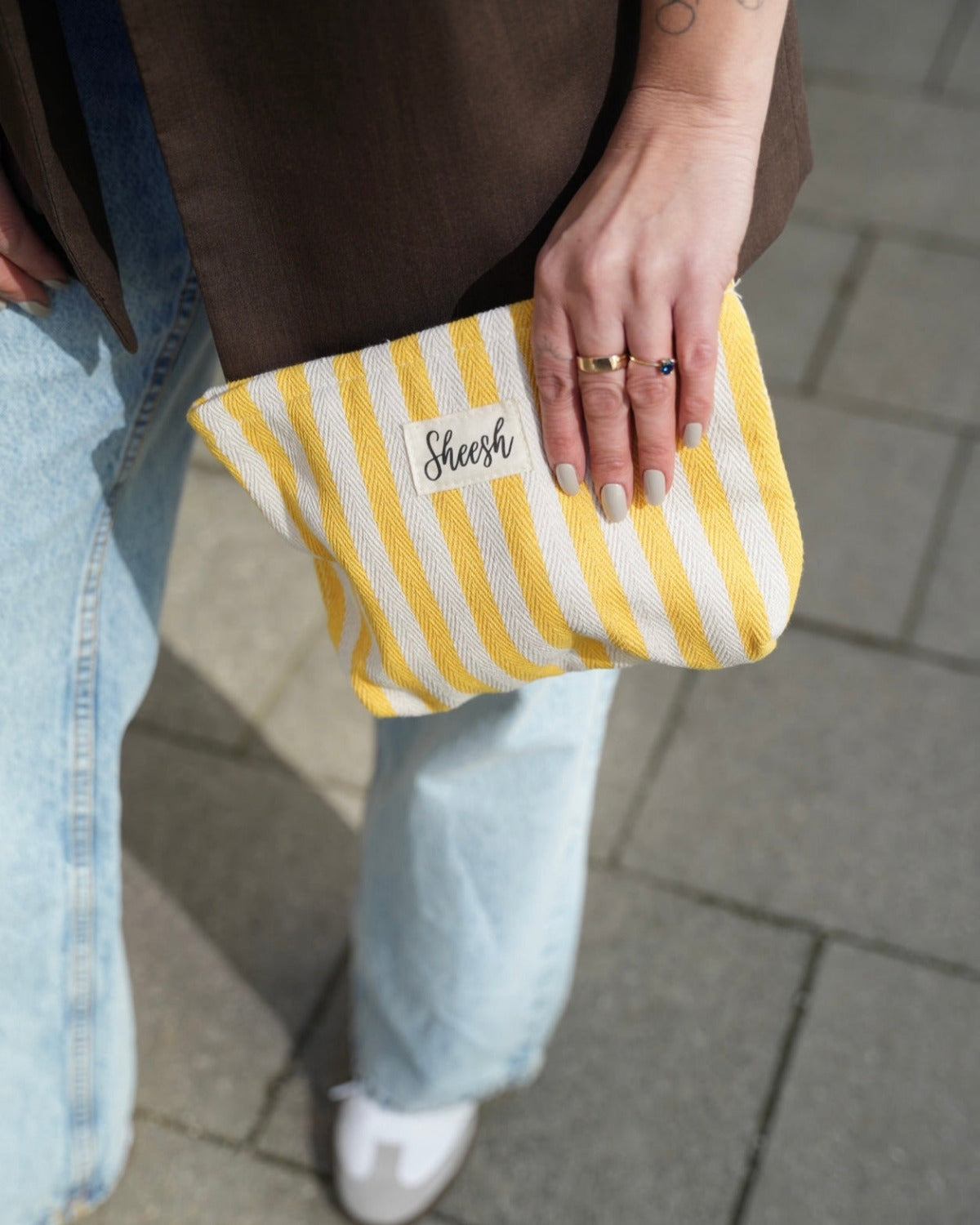 Detailaufnahme einer Hand, die eine gelb-weiß gestreifte Clutch-Tasche mit „Sheesh“-Logo hält. Die Person trägt eine braune Bluse, hellblaue Jeans und weiße Sneaker. Fokus auf der modernen Stofftasche als stylisches Accessoire für Frühling und Sommer, ideal für Mode-, Lifestyle- und Accessoire-Produktseiten von Sheesh Nails.