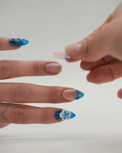 Auftragen von Sheesh Nails Press-on Nails in Blau mit zartem Blumenmotiv. Eine Hand setzt einen künstlichen Fingernagel präzise auf den natürlichen Nagel auf. Nahaufnahme des Anbringens von wiederverwendbaren Press-ons – einfache Anwendung, professionelle Optik und modernes Nageldesign für gepflegte Hände.