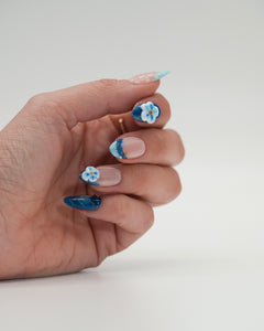 Sheesh Nails x PawsUp Press-on Nails in Blau und Hellblau mit 3D-Blüten und goldenen Details. Hand mit elegantem Nageldesign zeigt glänzende, wiederverwendbare künstliche Fingernägel im floralen Stil. Hochwertige Press-ons für professionelle Maniküre zu Hause – stilvolles Design mit Sommer-Vibes.