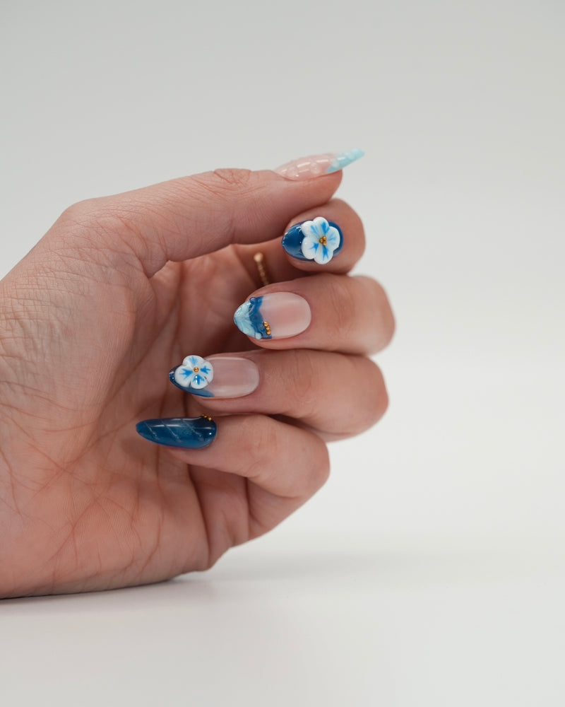 Sheesh Nails x PawsUp Press-on Nails in Blau und Hellblau mit 3D-Blüten und goldenen Details. Hand mit elegantem Nageldesign zeigt glänzende, wiederverwendbare künstliche Fingernägel im floralen Stil. Hochwertige Press-ons für professionelle Maniküre zu Hause – stilvolles Design mit Sommer-Vibes.