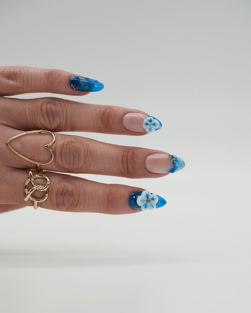 Sheesh Nails x PawsUp Press-on Nails in Blau mit transparentem Finish und 3D-Blüten. Hand mit goldenen Ringen zeigt glänzende, wiederverwendbare künstliche Fingernägel im modernen Design. Edles florales Press-on-Set für gepflegte Hände – stilvolle Maniküre mit Sommer- und Beach-Vibes.