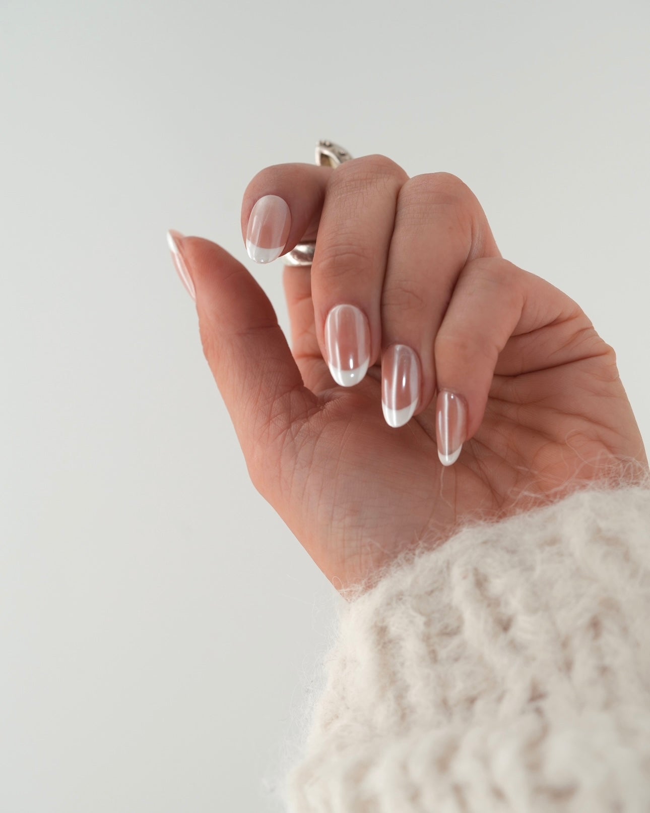 Sheesh Nails Press-on Nails in klassischem French-Design mit transparentem Glanzfinish. Hand mit cremefarbenem Strickpullover zeigt elegante, wiederverwendbare künstliche Fingernägel mit weißer French-Spitze. Zeitloses Nageldesign für gepflegte Hände – natürliche Eleganz und professionelle Maniküre für jeden Anlass.