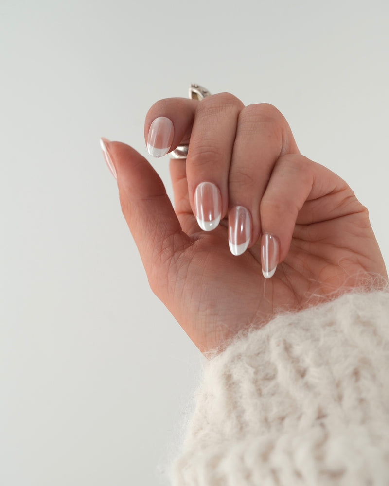 Sheesh Nails Press-on Nails in klassischem French-Design mit transparentem Glanzfinish. Hand mit cremefarbenem Strickpullover zeigt elegante, wiederverwendbare künstliche Fingernägel mit weißer French-Spitze. Zeitloses Nageldesign für gepflegte Hände – natürliche Eleganz und professionelle Maniküre für jeden Anlass.