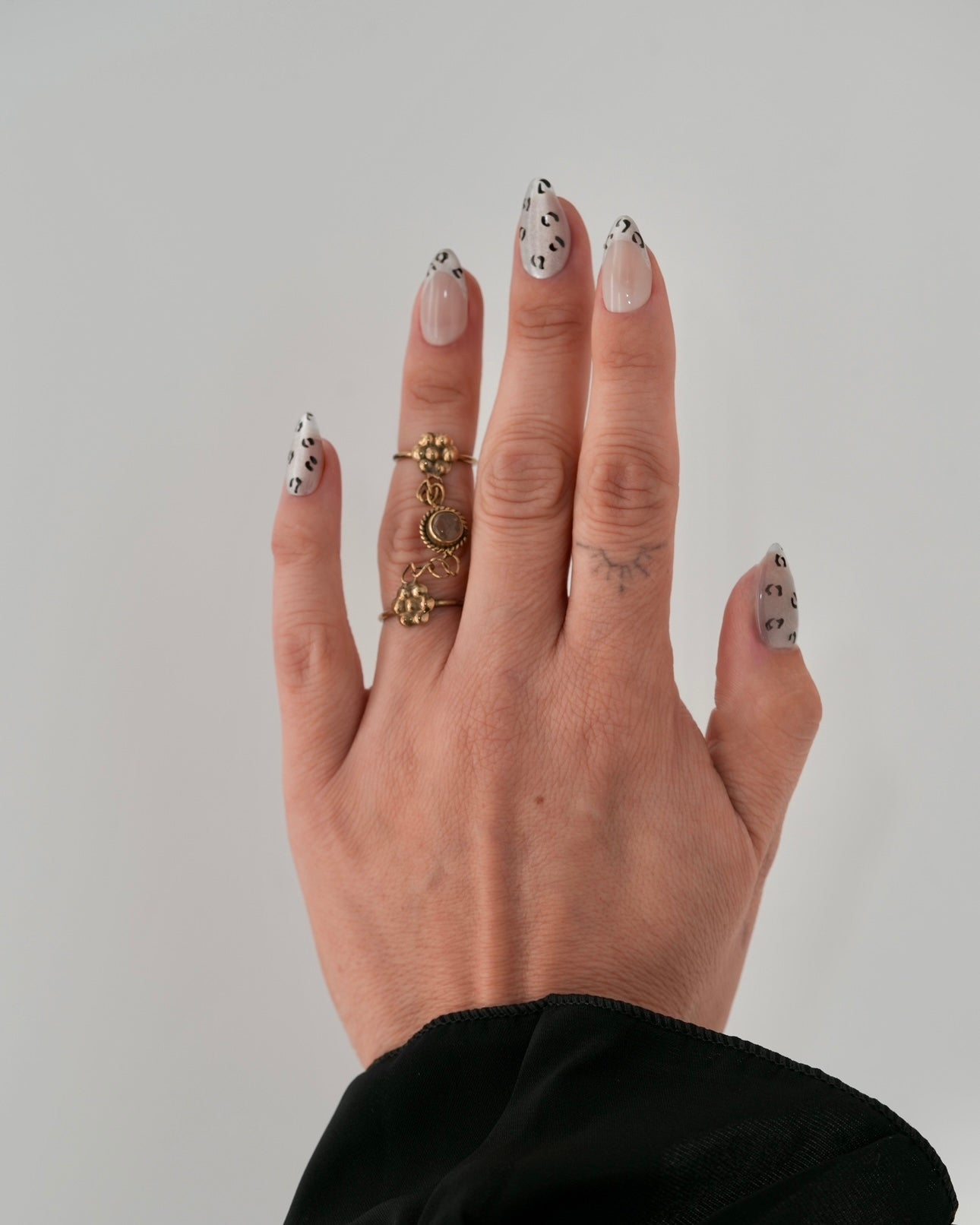 Sheesh Nails x PawsUp Press-on Nails mit schwarz-silbernem Leopardenmuster auf transparentem Untergrund. Hand mit goldenen Ringen präsentiert elegante, wiederverwendbare künstliche Fingernägel im modernen Animal-Print. Trendiges Press-on-Set für auffällige, stilvolle Maniküre mit Glamour-Faktor.