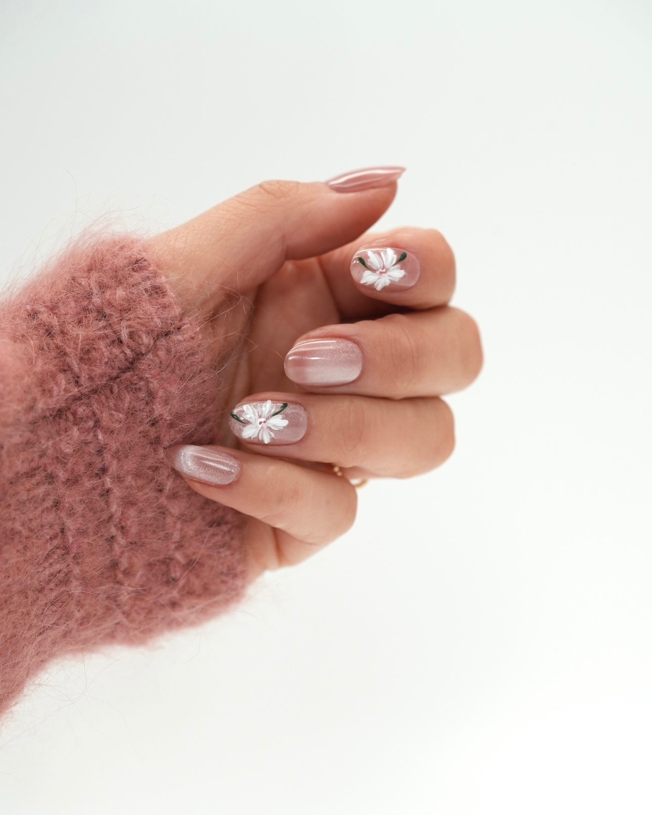 Sheesh Nails Press-on Nails in Rosé mit weißen Blüten und schimmerndem Glanzfinish. Hand in rosa Strickpullover zeigt elegante, wiederverwendbare künstliche Fingernägel im femininen Blumendesign. Hochwertige Press-ons für eine natürliche, gepflegte Maniküre mit edlem, romantischem Look.
