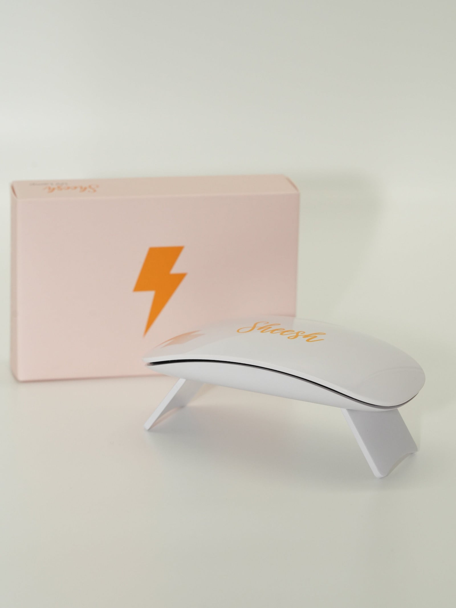 Weiße UV-Lampe mit orangefarbener „Sheesh“-Aufschrift steht vor einer rosafarbenen Box mit orangefarbenem Blitzsymbol. Das Produktfoto zeigt die Sheesh Nails UV-Lampe im schlichten, modernen Design – verwendet zum Aushärten der Nagelfolien.
