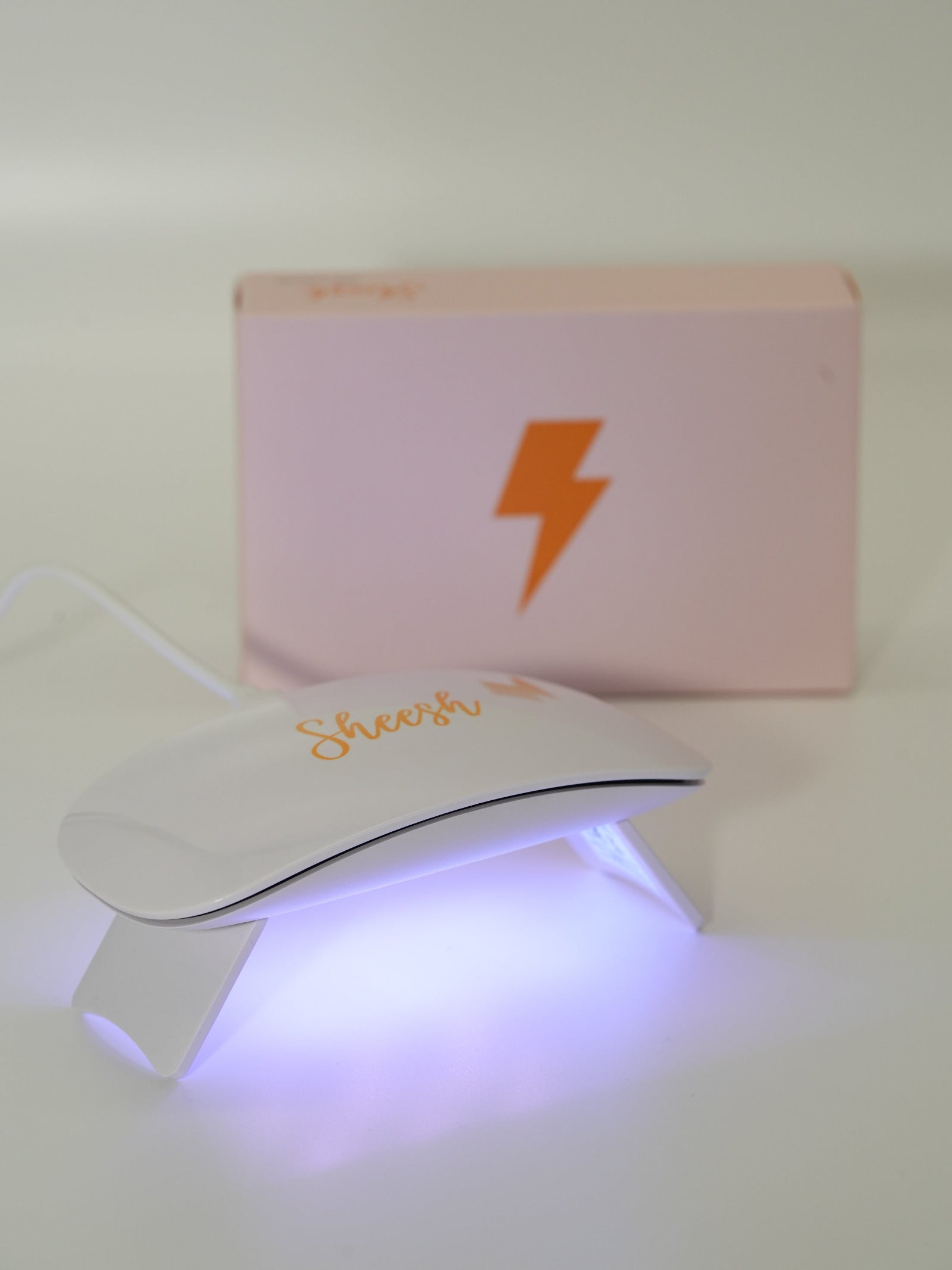 Weiße UV-Lampe mit goldener „Sheesh“-Aufschrift leuchtet in hellem Violettlicht, während sie in Betrieb ist. Dahinter steht eine rosafarbene Box mit goldfarbenem Blitzsymbol. Das Bild zeigt die Sheesh Nails UV-Lampe beim Aushärten der Nagelfolien.
