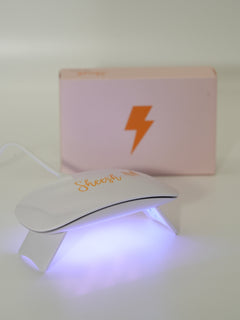 Weiße UV-Lampe mit goldener „Sheesh“-Aufschrift leuchtet in hellem Violettlicht, während sie in Betrieb ist. Dahinter steht eine rosafarbene Box mit goldfarbenem Blitzsymbol. Das Bild zeigt die Sheesh Nails UV-Lampe beim Aushärten der Nagelfolien.