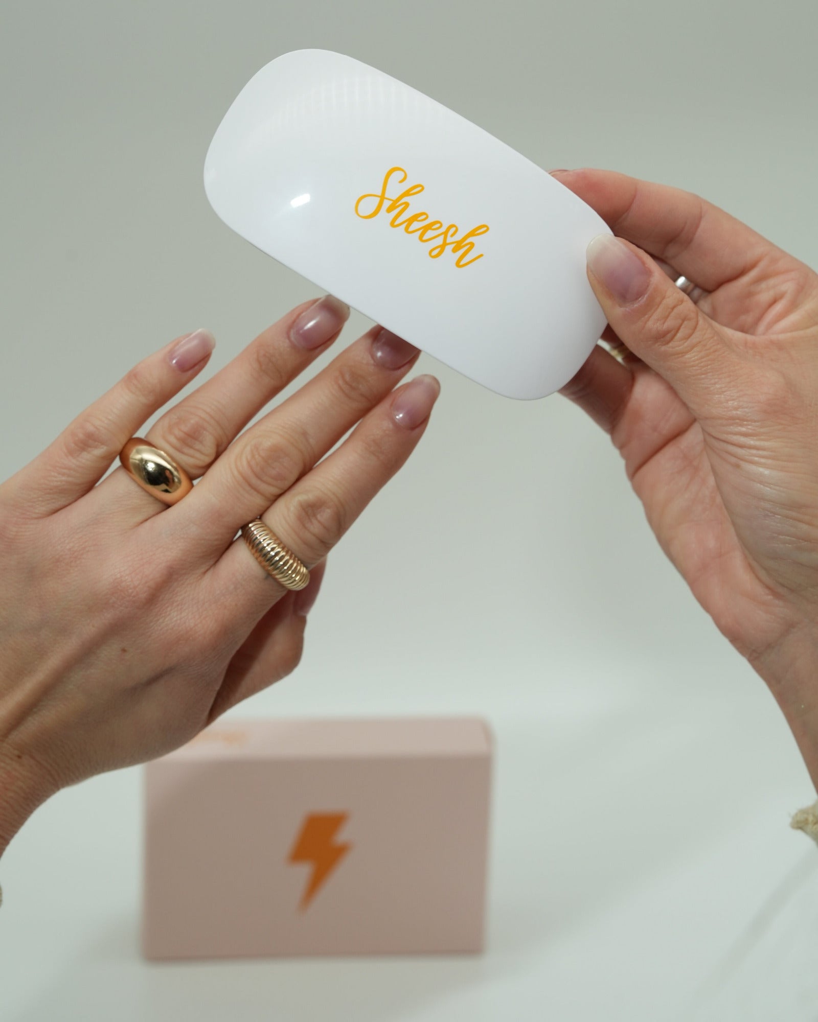 Hand mit natürlich gepflegten Nägeln hält eine weiße UV-Lampe mit goldener „Sheesh“-Aufschrift. Im Hintergrund steht eine rosafarbene Box mit Blitzsymbol. Das Bild zeigt die Sheesh Nails UV-Lampe beim Einsatz zum Aushärten der Nagelfolien.