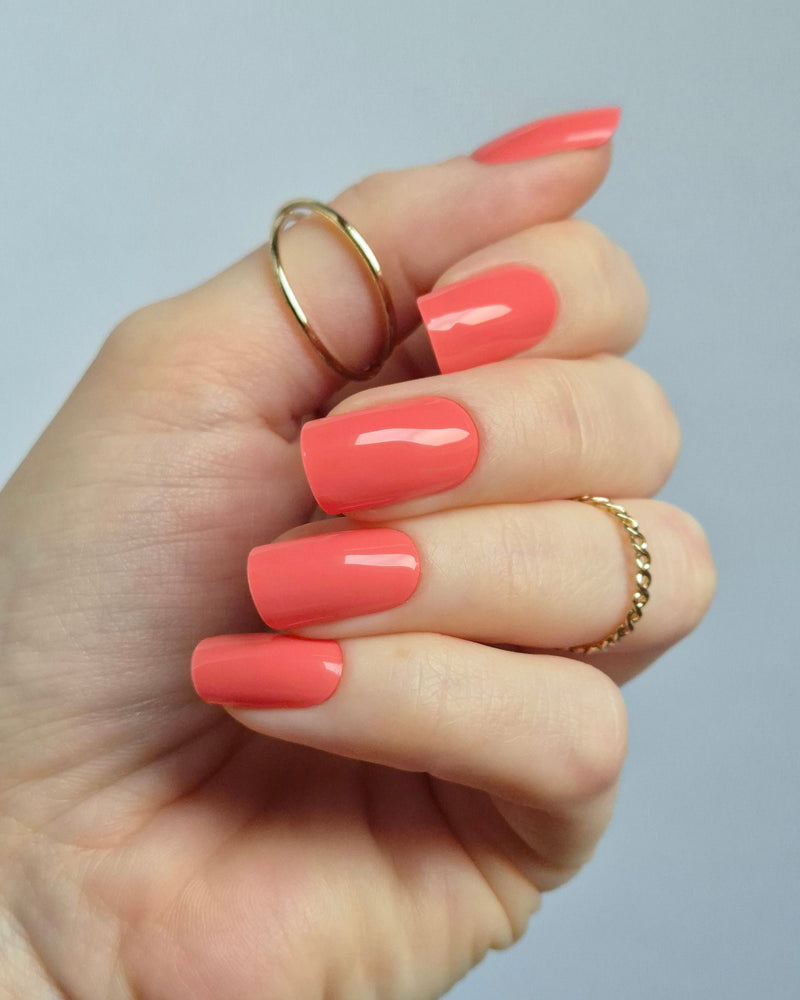 Hand mit korallroter UV-Maniküre von SHEESH Nails – „Lobster Roll“. Glänzende, gleichmäßig aufgetragene Gel-Nagelfolien in intensivem Korallton. Vegane, selbstklebende und lang haltbare Maniküre für ein sommerlich frisches Nail-Design zuhause.