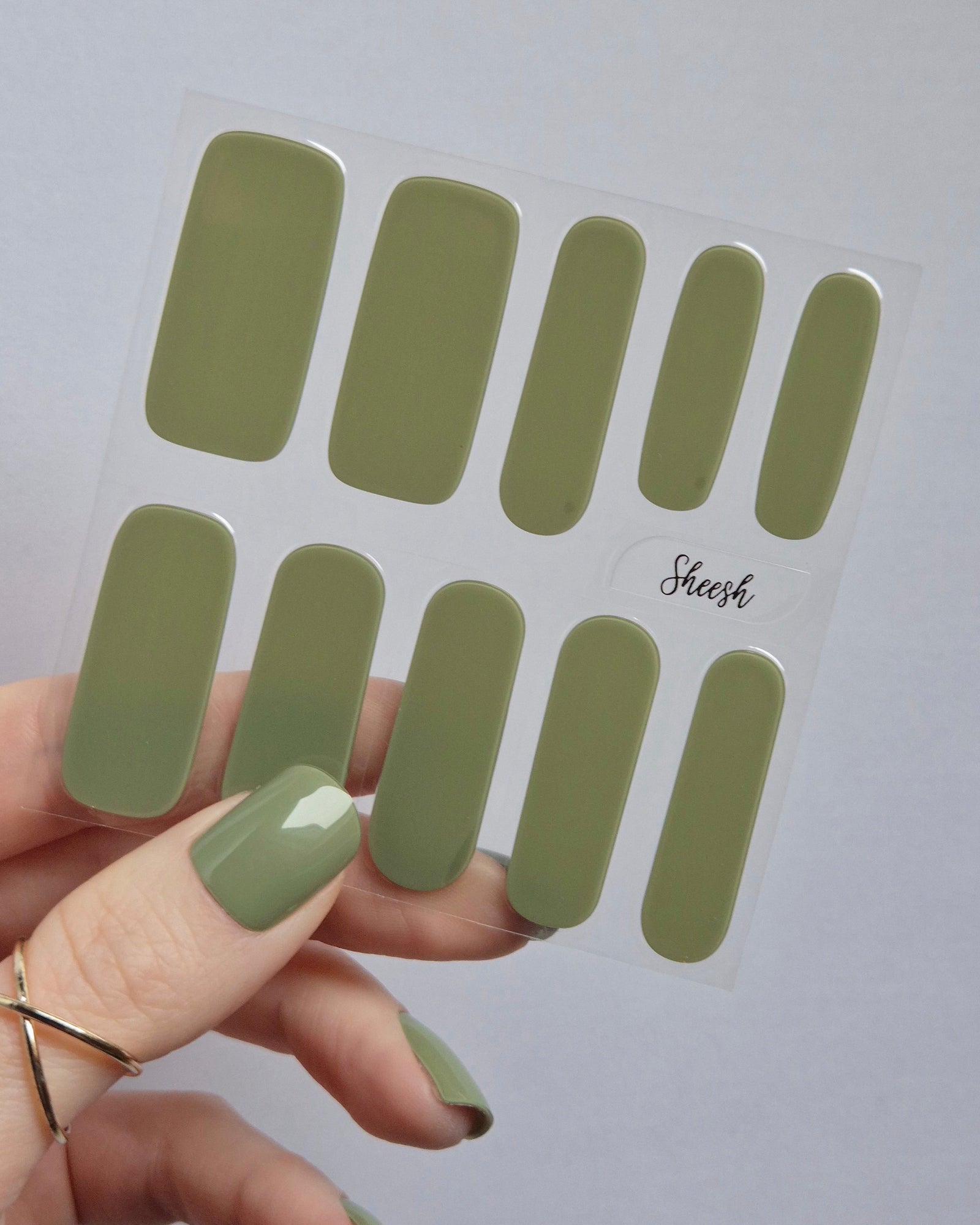 UV Nagelfolien in Olivgrün – SHEESH Nails „Olives from Napa“. Hand hält glänzende olivgrüne Gel-Nagelfolien auf transparenter Trägerfolie. Perfekte Maniküre ohne Lack, vegan, lang haltbar und einfach anzuwenden.