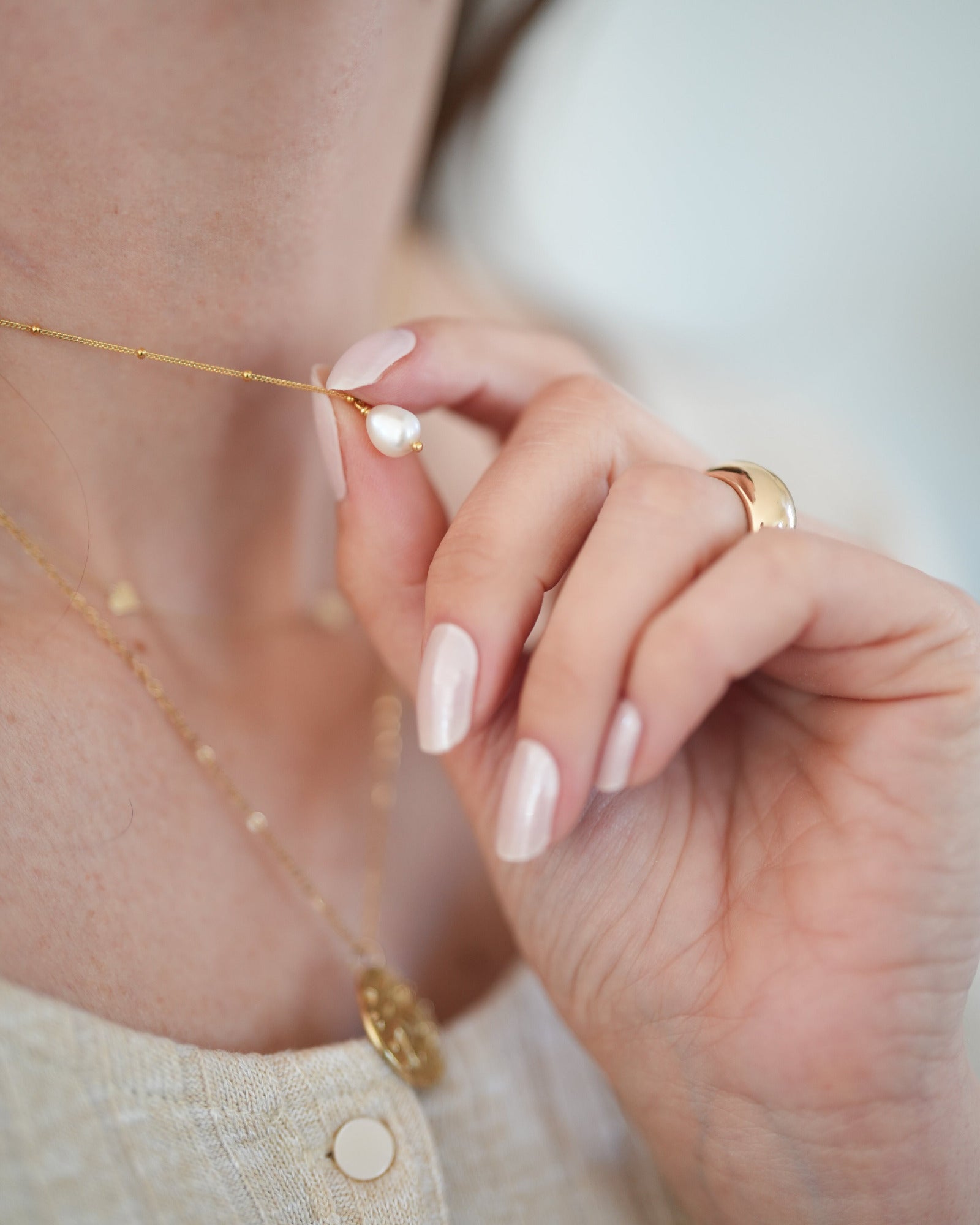 Hand mit perlmuttfarben lackierten Nägeln hält eine goldene Halskette mit Perlenanhänger. Die Nägel glänzen dezent und wirken elegant – getragen wird die UV-Nagelfolie „Pearl Diver“ von Sheesh Nails.