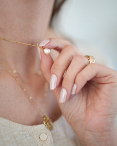 Hand mit perlmuttfarben lackierten Nägeln hält eine goldene Halskette mit Perlenanhänger. Die Nägel glänzen dezent und wirken elegant – getragen wird die UV-Nagelfolie „Pearl Diver“ von Sheesh Nails.