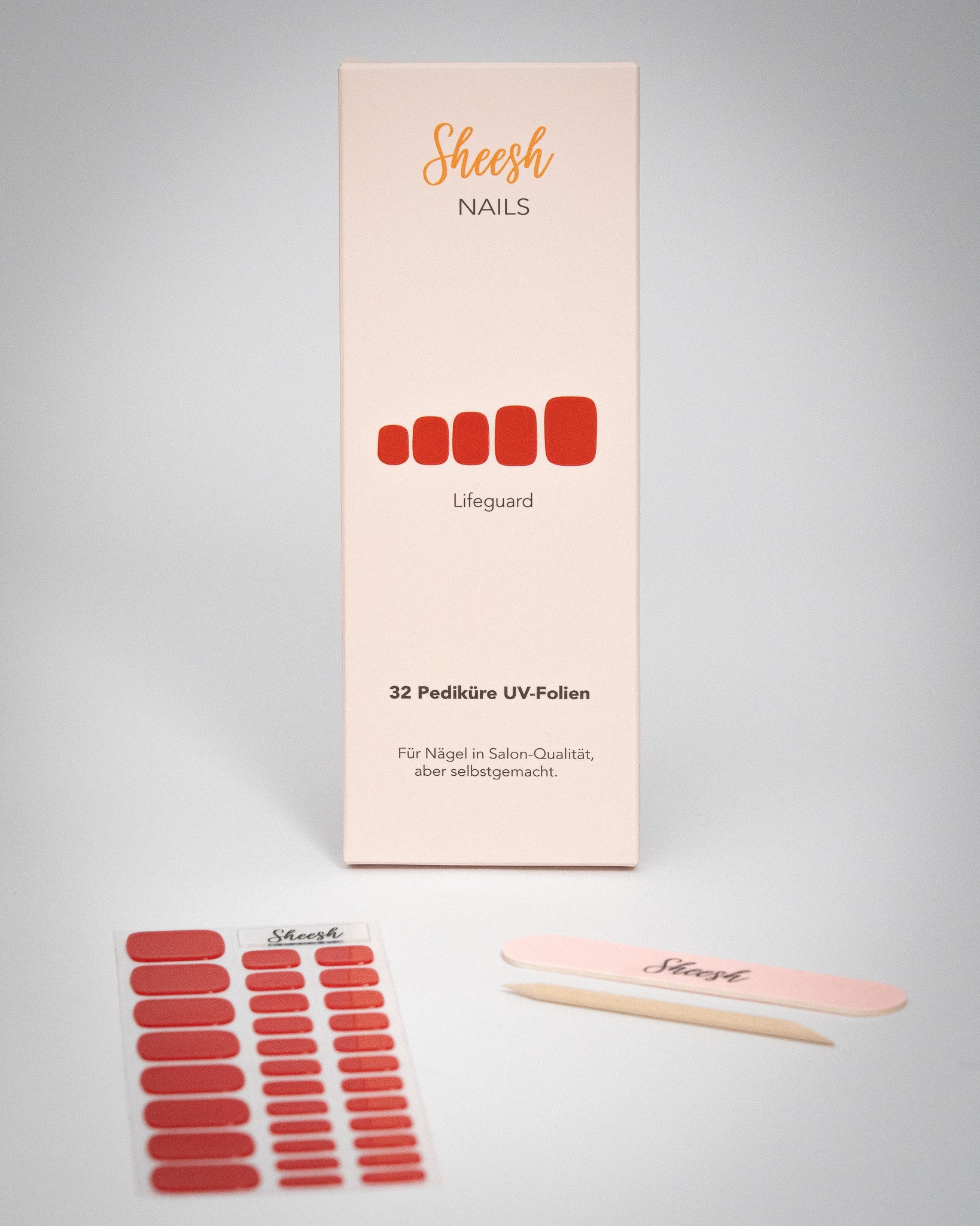 Produktaufnahme der Sheesh Nails Pedi UV-Nagelfolie „Lifeguard“. Die Verpackung präsentiert das kräftige Rot der Folien, kombiniert mit Feile und Holzstäbchen. Das minimalistische Setup und das helle Licht betonen die hochwertige Qualität und den modernen, gepflegten Salon-Look – ideal für elegante Sommerpediküren mit leuchtendem Finish.