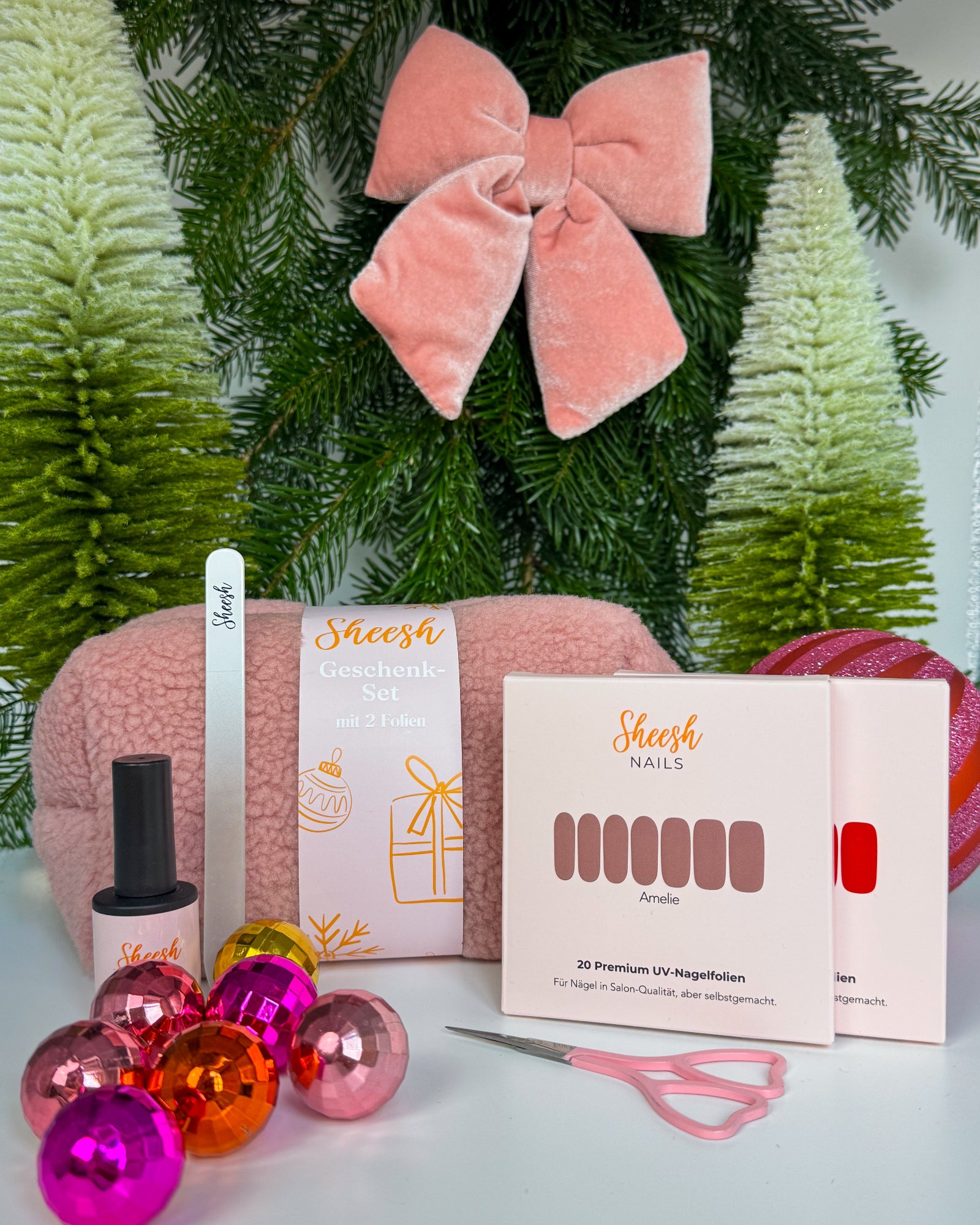 Weihnachtlich dekoriertes Set mit Sheesh Nails Nagelfolien, Topcoat, Glasfeile, Schere und flauschiger rosa Teddytasche. Im Hintergrund Tannenzweige, Christbaumkugeln und eine rosa Samtschleife. Das Weihnachtsgeschenke-Set von Sheesh Nails ist ideal als Beauty-Geschenk für gepflegte Nägel und festliche Maniküre.