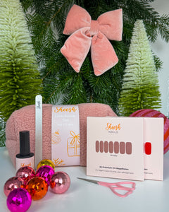 Weihnachtlich dekoriertes Set mit Sheesh Nails Nagelfolien, Topcoat, Glasfeile, Schere und flauschiger rosa Teddytasche. Im Hintergrund Tannenzweige, Christbaumkugeln und eine rosa Samtschleife. Das Weihnachtsgeschenke-Set von Sheesh Nails ist ideal als Beauty-Geschenk für gepflegte Nägel und festliche Maniküre.