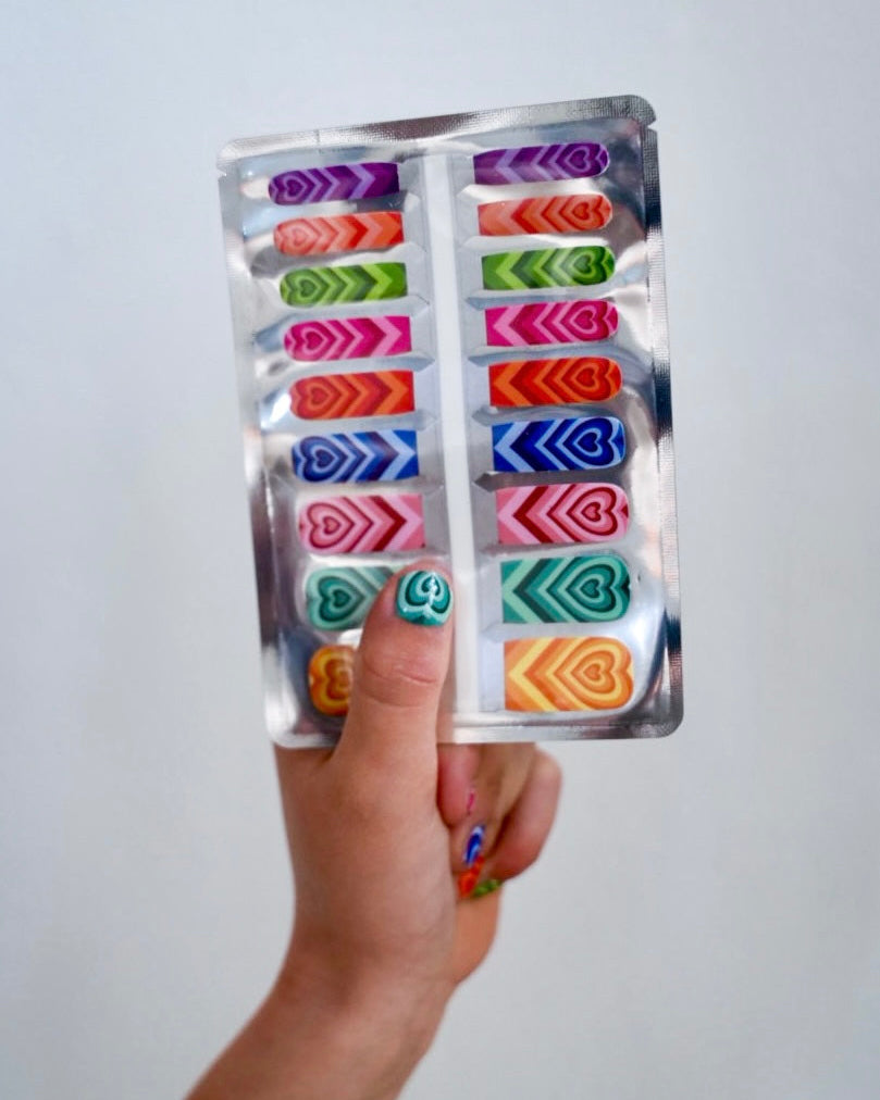 Nahaufnahme einer Kinderhand mit Sheesh Nails Kinder-Nagelfolien „Herzi“. Die selbstklebenden Sticker zeigen farbenfrohe Herzmuster in kräftigen Regenbogenfarben. Das verspielte Design sorgt für einen fröhlichen, kindgerechten Look. Die Folien sind hautfreundlich, ungiftig und leicht anzubringen – ideal für kreative Kinder-Maniküren mit Farbe und Spaß.