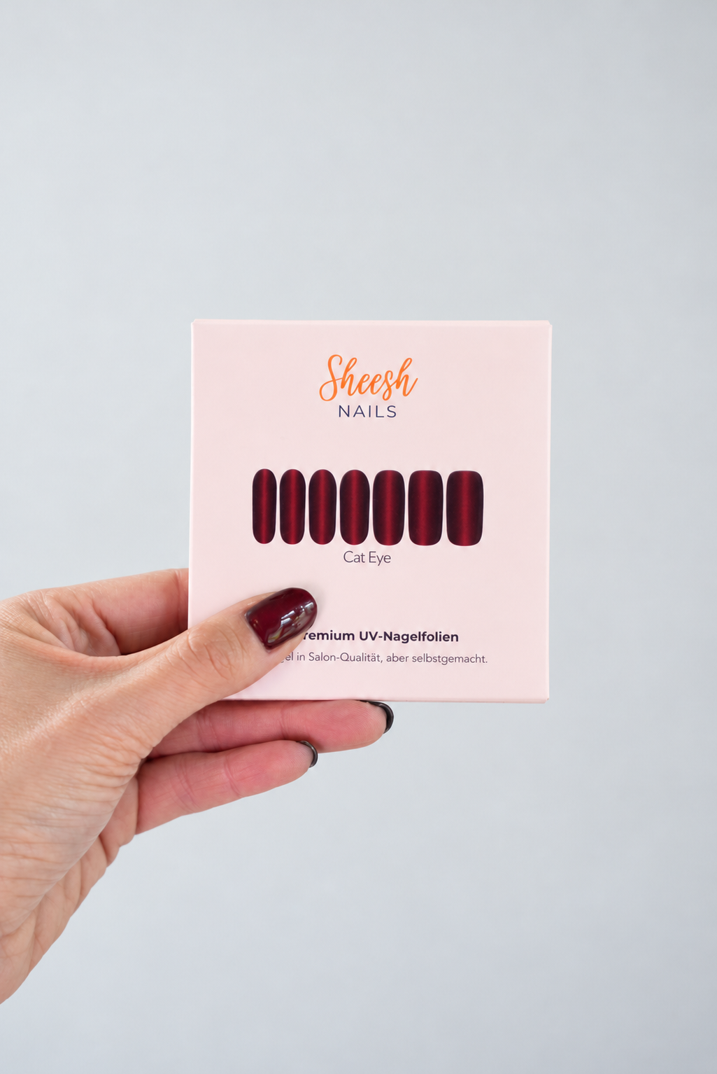 SHEESH Nails – Cat Eye Verpackung in einer Hand mit bordeauxroter Maniküre.