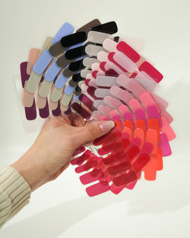 Produktfoto eines Folienfächers mit verschiedenen Sheesh Nails UV-Nagelfolien in zahlreichen Farben. Die Hand hält den aufgefächerten Überblick von Rot-, Rosa-, Lila- und Blautönen. Ideal zur Farbauswahl und Präsentation des Sortiments – professionell, übersichtlich und ästhetisch arrangiert.