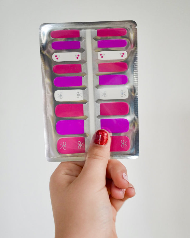 Kinderhand hält SHEESH Nails Nagelfolien Cherry Bow in Pink und Weiß mit kleinen Schleifen- und Kirschenmotiven – süßes Nageldesign für Kinder.