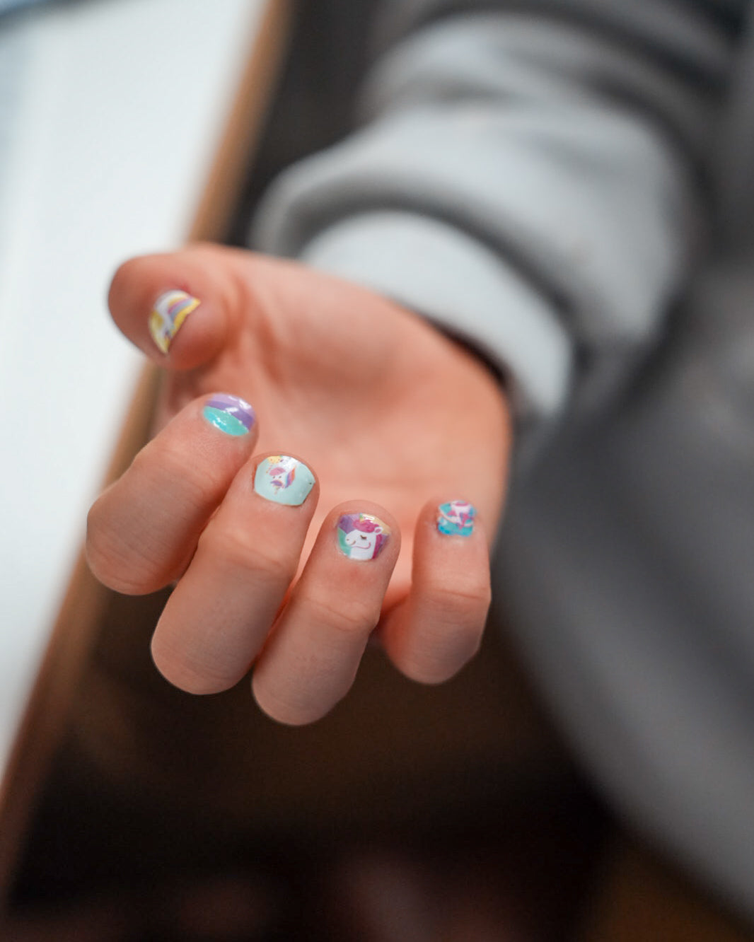 Nahaufnahme einer Kinderhand mit den Sheesh Nails Kinder-Nagelfolien „Einhorn“. Die selbstklebenden Sticker zeigen fröhliche Einhorn-Motive auf pastellfarbenem Hintergrund mit Regenbögen und Sternen. Das deckende Finish sorgt für einen bunten, verspielten Look. Hautfreundlich, ungiftig und leicht aufzubringen – ideal für kreative Kinder-Maniküren mit Magie und Fantasie.