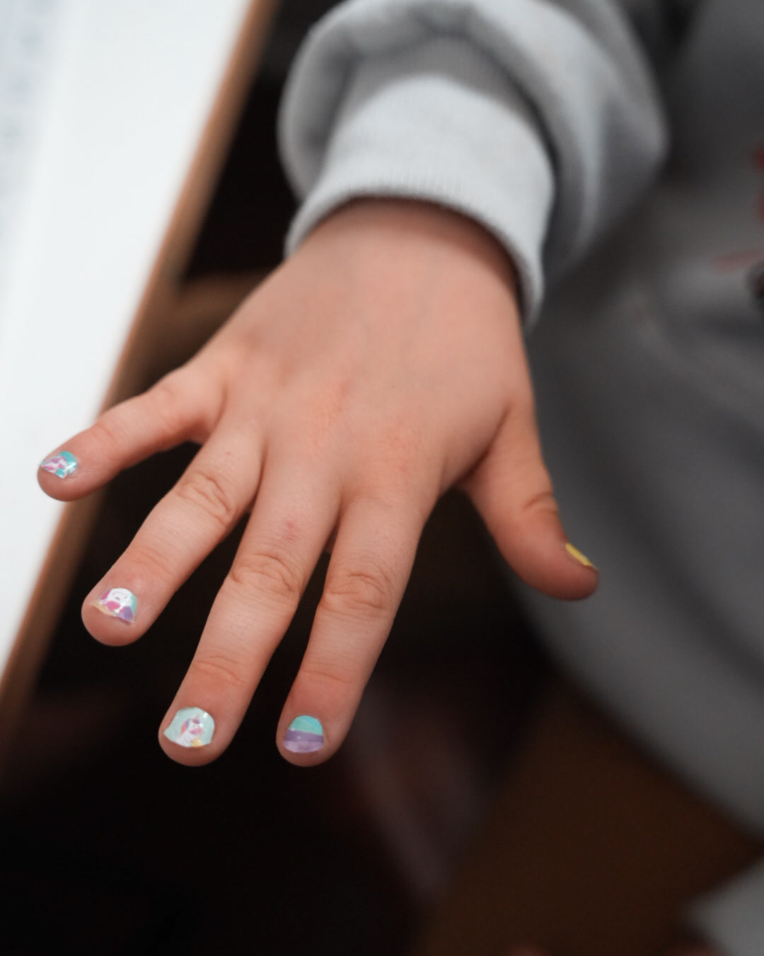 Nahaufnahme einer Kinderhand mit den Sheesh Nails Kinder-Nagelfolien „Einhorn“. Die selbstklebenden Sticker zeigen fröhliche Einhorn-Motive auf pastellfarbenem Hintergrund mit Regenbögen und Sternen. Das deckende Finish sorgt für einen bunten, verspielten Look. Hautfreundlich, ungiftig und leicht aufzubringen – ideal für kreative Kinder-Maniküren mit Magie und Fantasie.
