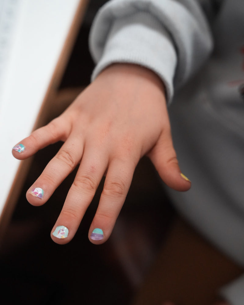 Nahaufnahme einer Kinderhand mit den Sheesh Nails Kinder-Nagelfolien „Einhorn“. Die selbstklebenden Sticker zeigen fröhliche Einhorn-Motive auf pastellfarbenem Hintergrund mit Regenbögen und Sternen. Das deckende Finish sorgt für einen bunten, verspielten Look. Hautfreundlich, ungiftig und leicht aufzubringen – ideal für kreative Kinder-Maniküren mit Magie und Fantasie.