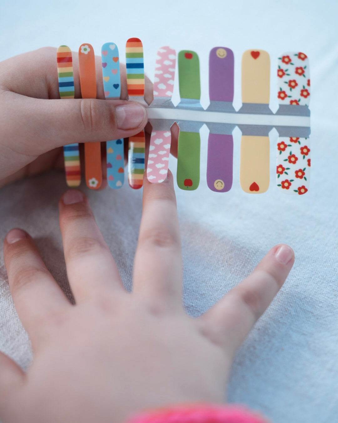 Nahaufnahme einer Kinderhand mit den Sheesh Nails Kinder-Nagelfolien „Happy“. Die bunten, selbstklebenden Sticker zeigen fröhliche Muster in Regenbogenfarben, Herzen und Punkten – perfekt für kleine Nail-Artists, die Spaß an Farbe und Kreativität haben. Die Sticker sind deckend, leicht aufzubringen und rückstandsfrei wieder zu entfernen – ganz ohne UV-Lampe. Hautfreundlich, ungiftig und ideal für kreative Kinder-Maniküren voller Freude und Fantasie.