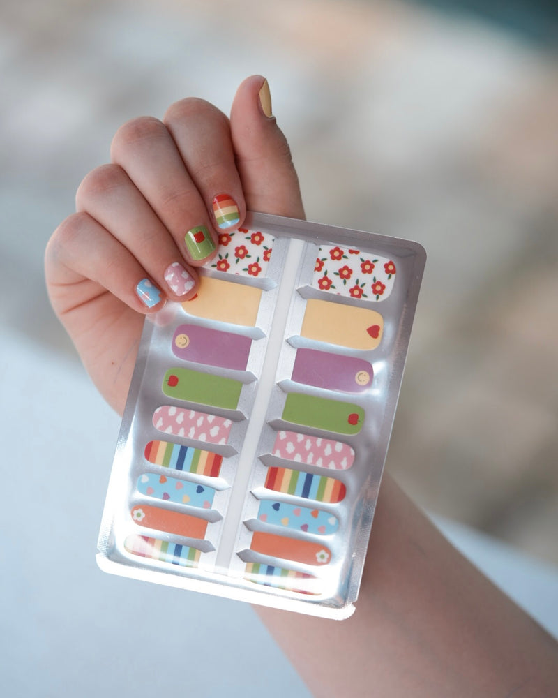 Nahaufnahme einer Kinderhand mit den Sheesh Nails Kinder-Nagelfolien „Happy“. Die bunten, selbstklebenden Sticker zeigen fröhliche Muster in Regenbogenfarben, Herzen und Punkten – perfekt für kleine Nail-Artists, die Spaß an Farbe und Kreativität haben. Die Sticker sind deckend, leicht aufzubringen und rückstandsfrei wieder zu entfernen – ganz ohne UV-Lampe. Hautfreundlich, ungiftig und ideal für kreative Kinder-Maniküren voller Freude und Fantasie.