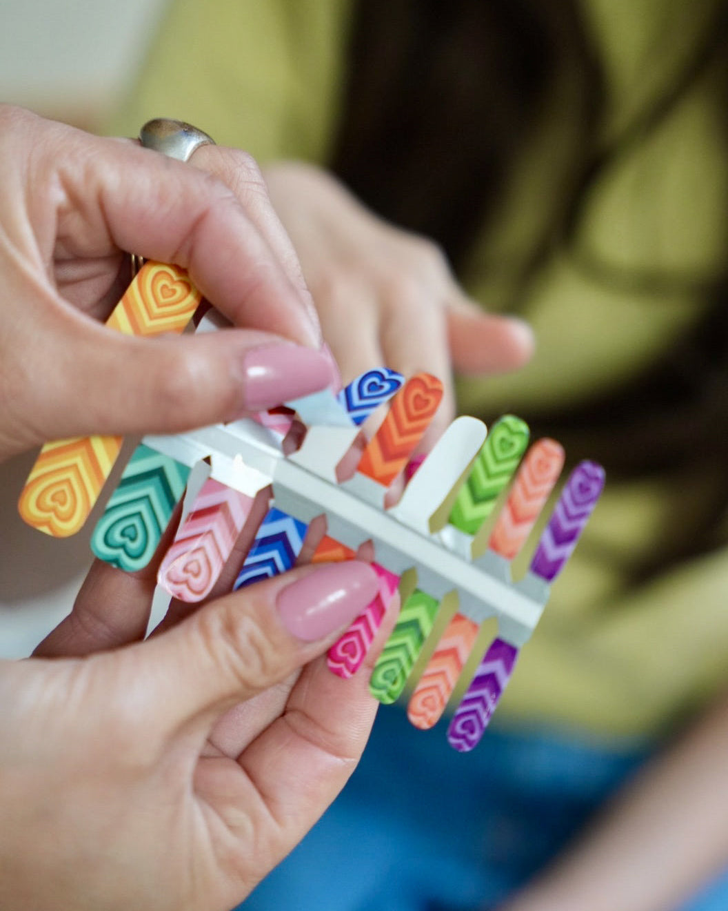 Nahaufnahme beim Zuschneiden einer Sheesh Nails Kinder-Nagelfolie „Herzi“. Eine erwachsene Hand hilft einem Kind beim Anpassen der Folie mit buntem Herzmotiv. Das detailreiche Design ist farbenfroh und verspielt. Die Folien sind hautfreundlich, ungiftig und einfach anzubringen – ideal für kreative Kinder-Maniküren mit Herzmotiv.