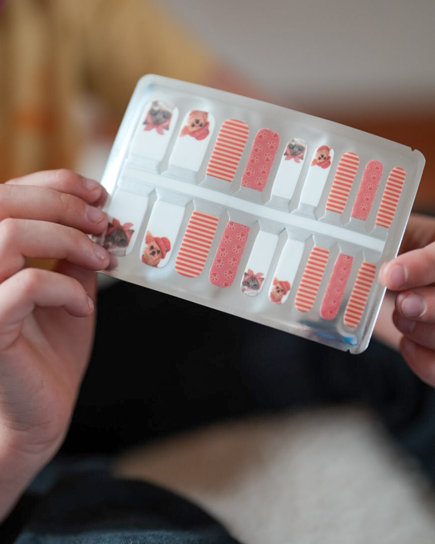 Nahaufnahme einer Kinderhand, die die Sheesh Nails Kinder-Nagelfolien „Hot Dog“ in der Verpackung hält. Die selbstklebenden Sticker zeigen ein fröhliches Design in Orange mit Streifen und kleinen Hundemotiven. Das deckende Finish sorgt für einen farbenfrohen, kindgerechten Look. Hautfreundlich, ungiftig und einfach anzubringen – ideal für kreative Kinder-Maniküren mit Spaß und Fantasie.