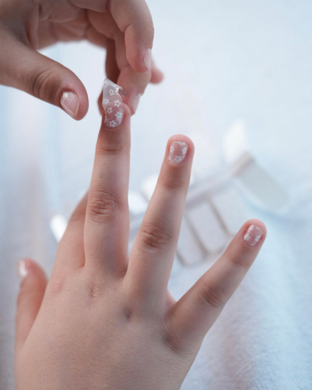 Nahaufnahme einer Kinderhand mit den Sheesh Nails Kinder-Nagelfolien „Lilly“. Die transparenten Sticker zeigen feine, weiße Blumen und lassen die natürliche Nagelfarbe sanft durchscheinen. Ideal für kleine Nail-Artists, die verspielte, zarte Designs lieben. Die selbstklebenden Folien sind leicht anzubringen, hautfreundlich, ungiftig und lassen sich rückstandsfrei wieder entfernen – ganz ohne UV-Lampe. Perfekt für kreative Kinder-Maniküren mit einem natürlichen und liebevollen Look.